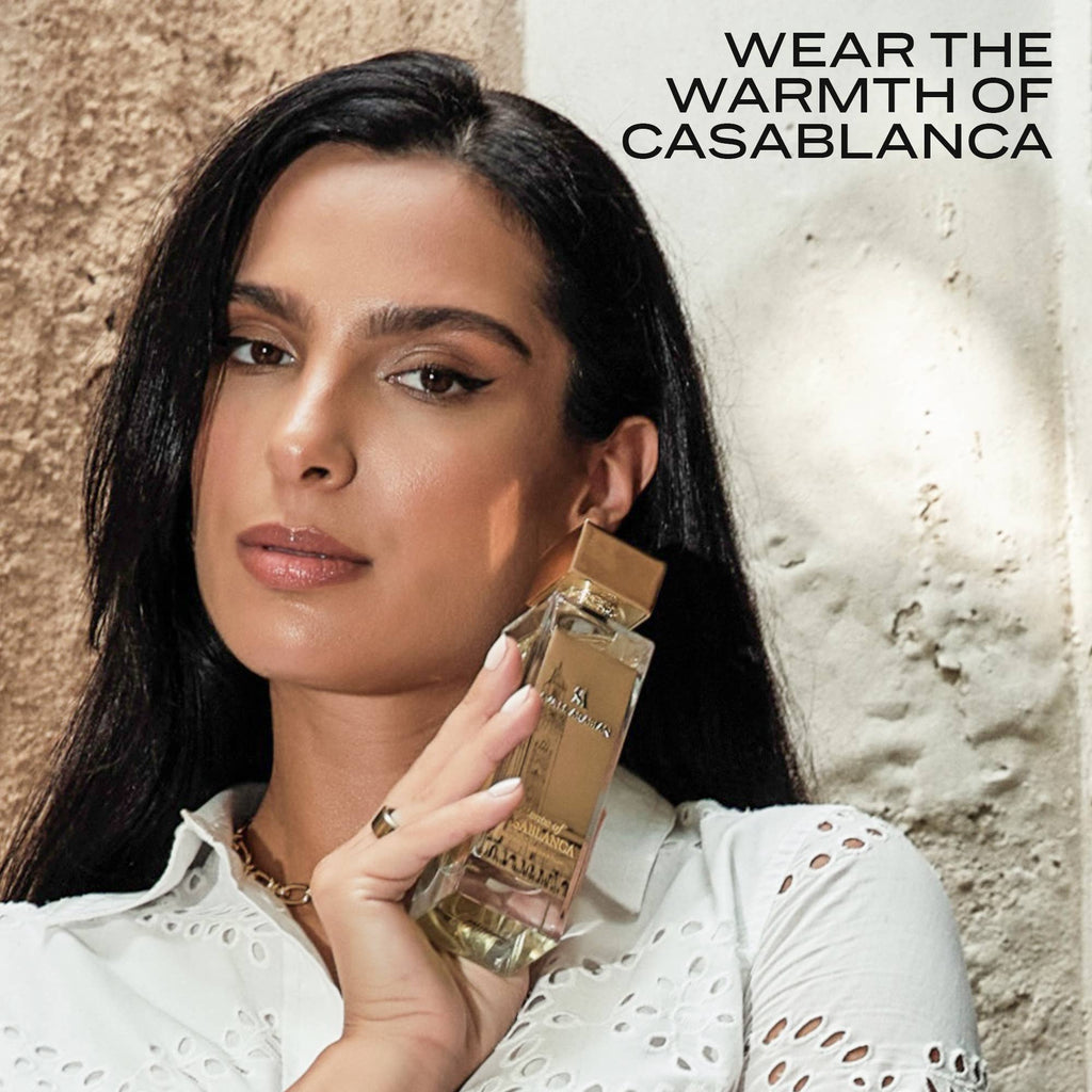 Woman in a white eyelet blouse holding the Swiss Arabian Essence of Casablanca bottle against a sunlit plaster wall, captioned Wear The Warmth Of Casablanca